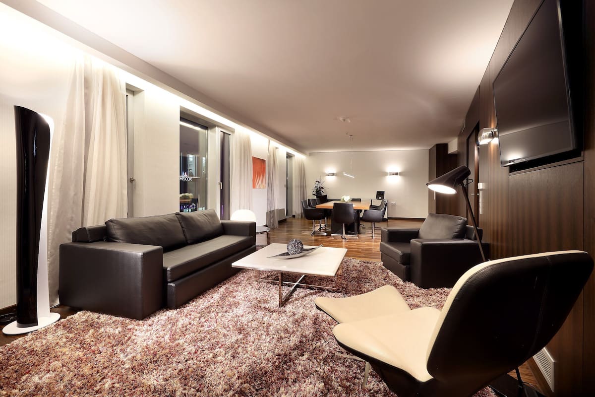 A modern living room with a sofa, coffee table, and chairs. There is a flat-screen TV mounted on the wall and a dining area in the background.