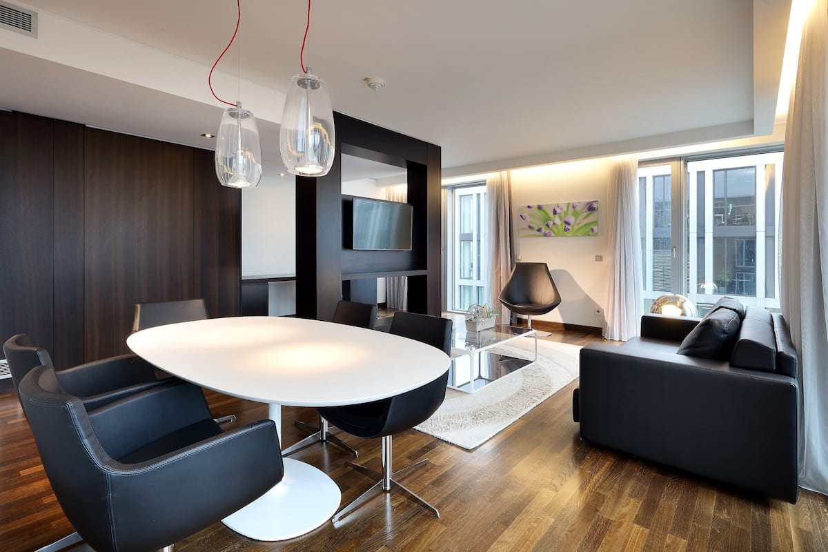 A modern living room with a round dining table, black leather chairs, a black sofa, and a TV mounted on the wall.
