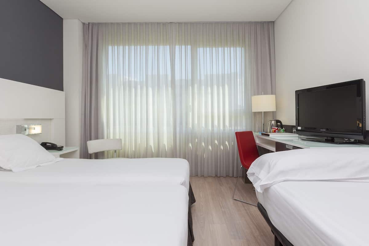Hotellihuone, jossa on kaksi sänkyä, työpöytä, jossa on televisio, punainen tuoli ja ikkuna, jossa on verhot.