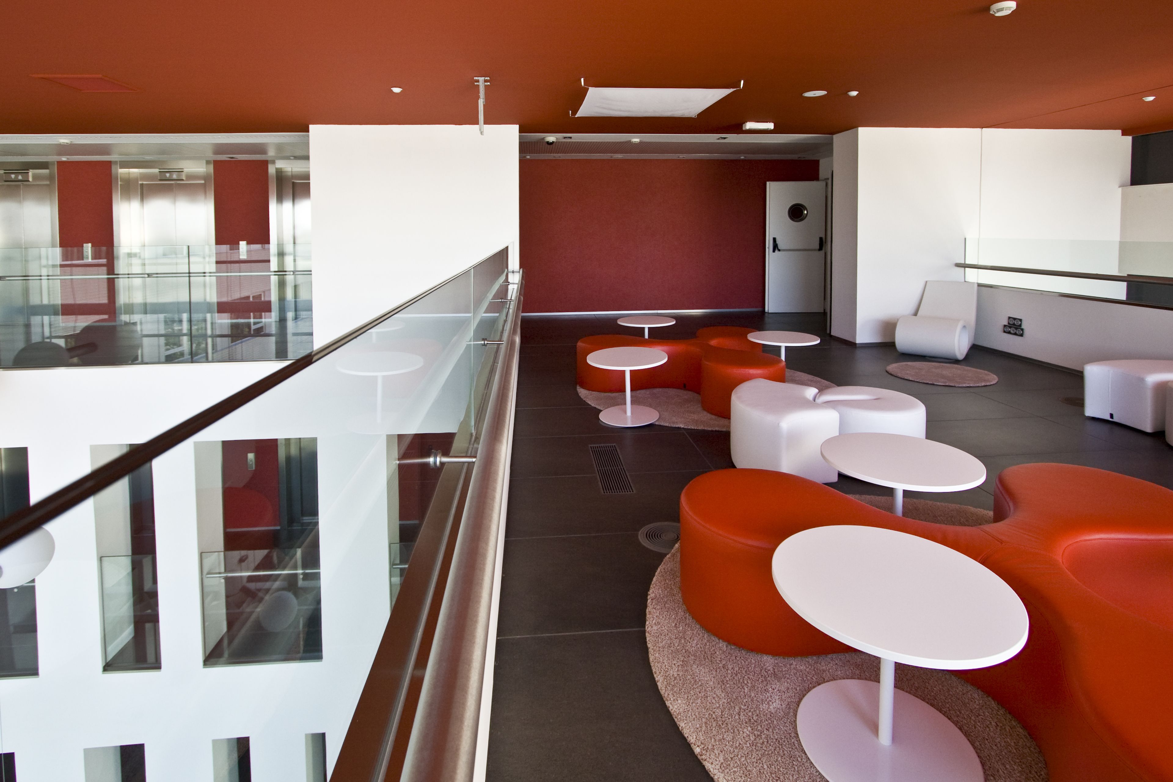 A modern lounge area with glass railings, orange and white seating, and a view of the interior space.