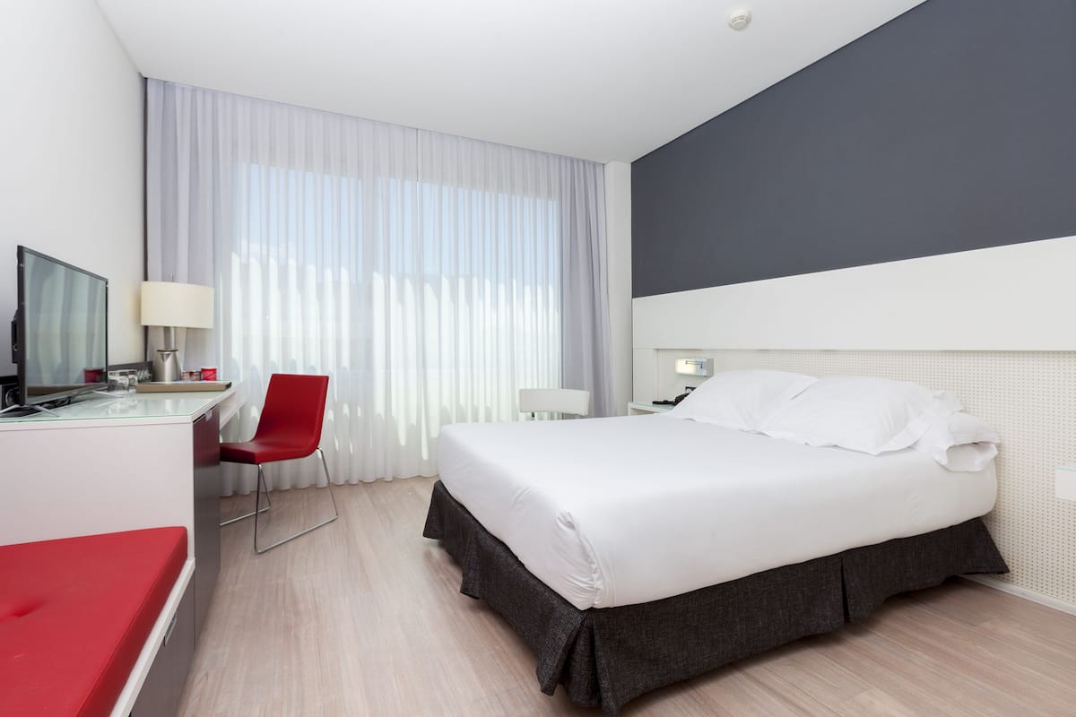 Hotellihuone, jossa on sänky, työpöytä tuolilla, televisio ja ikkuna, jossa on verhot.