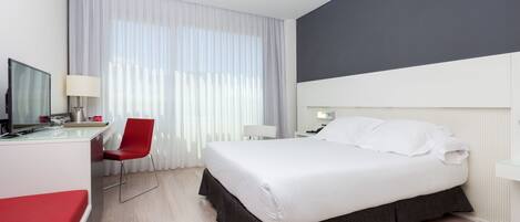 A hotel room with a bed, a desk with a chair, a TV, and a window with curtains.