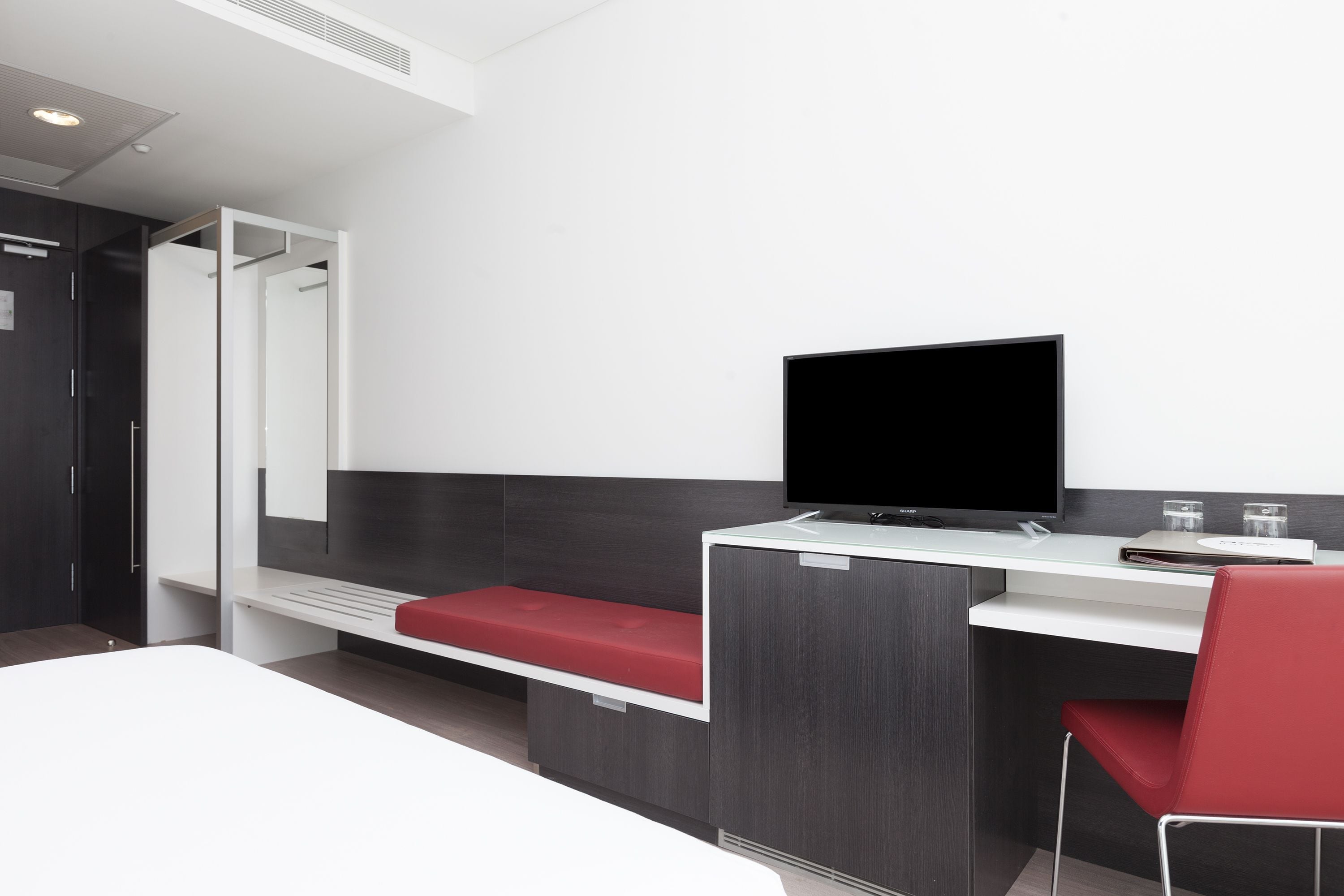 Quarto de hotel moderno com uma cama, uma escrivaninha com um computador, uma cadeira vermelha e um espelho grande.