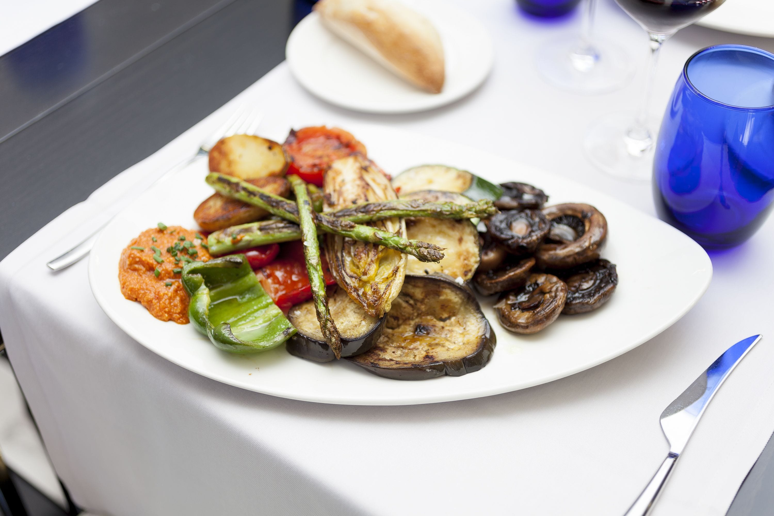 A plate of roasted vegetables including asparagus, tomatoes, and mushrooms, served on a white plate with a fork and knife beside it.