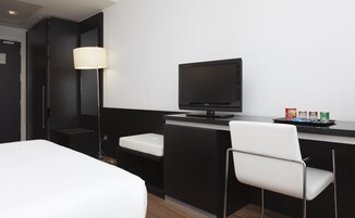 Ein modernes Hotelzimmer mit Bett, Schreibtisch mit Computer, Stuhl, Lampe und einer kleinen Minibar.
