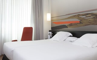 Ein Hotelzimmer mit Bett, einem roten Sessel, einem Bild an der Wand und einem Fenster mit Vorhängen.