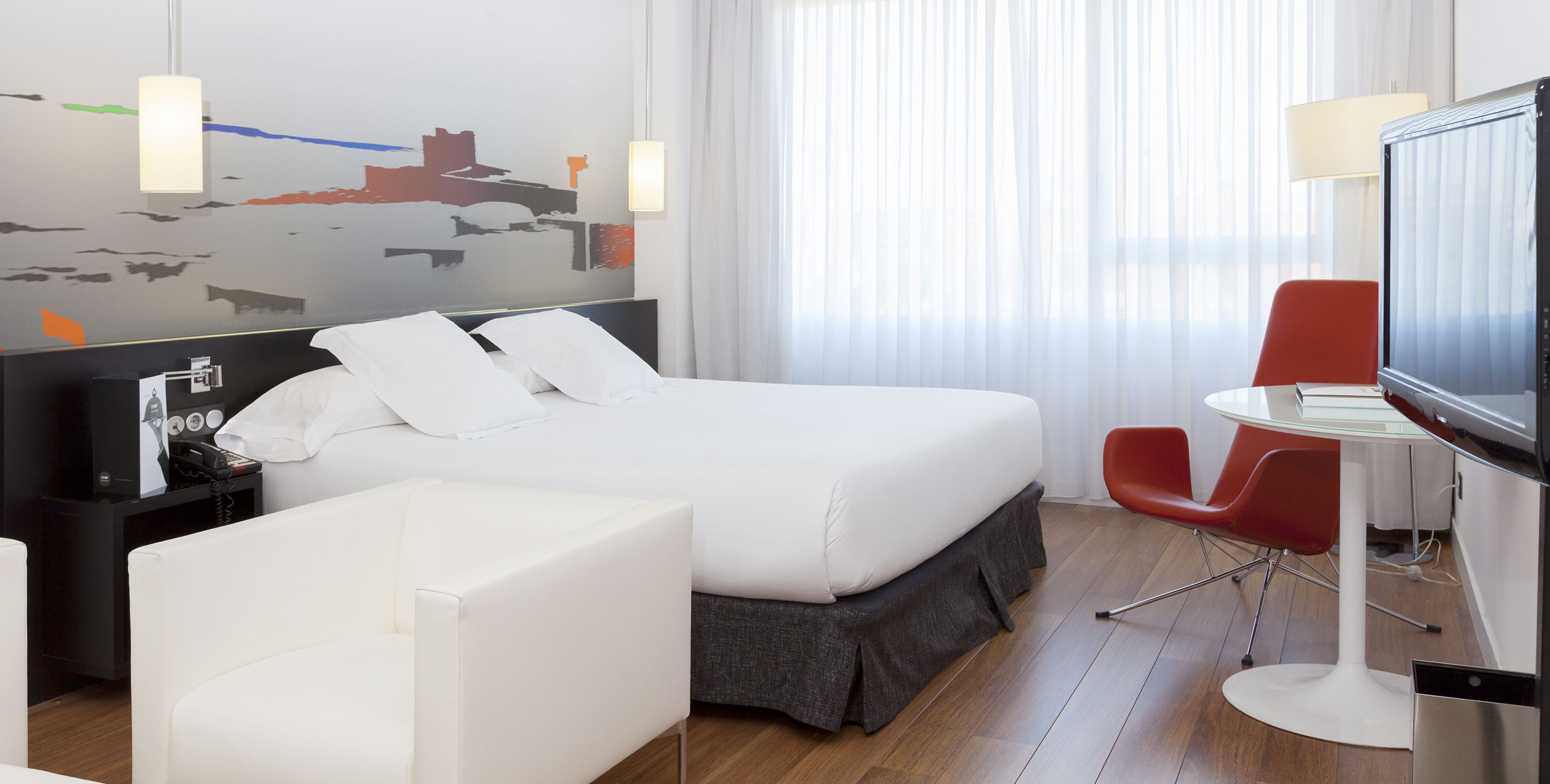 Une chambre d’hôtel moderne avec un grand lit, un bureau avec une télévision, un fauteuil rouge et une fresque représentant un paysage urbain sur le mur.