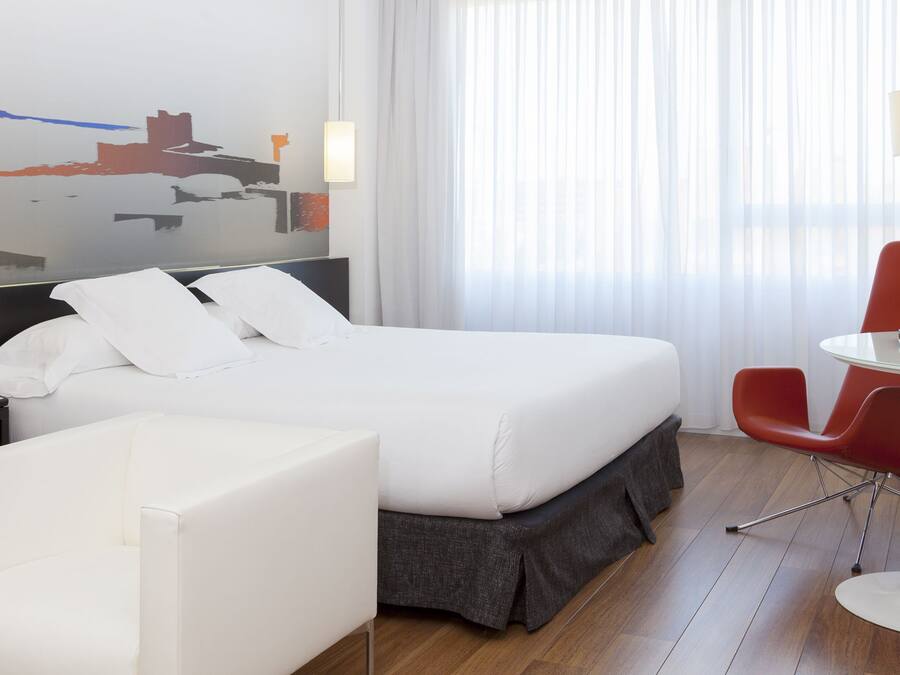 Une chambre d’hôtel moderne avec un grand lit, un bureau avec une télévision, un fauteuil rouge et une fresque représentant un paysage urbain sur le mur.