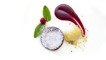 Un postre con base de chocolate, una bola de helado de vainilla y adornos que incluyen una baya roja, hojas de menta y azúcar glas.