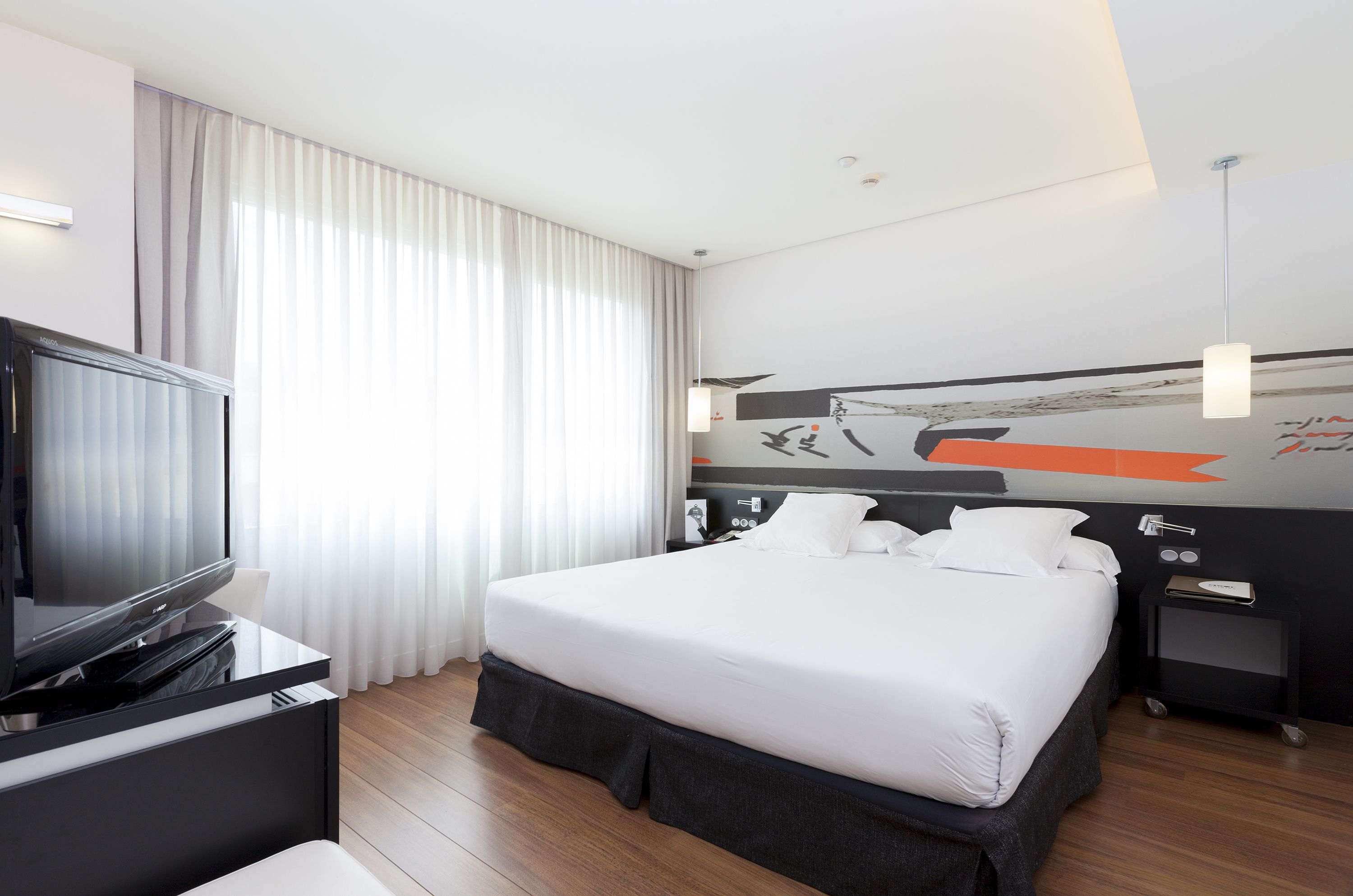 Una habitación de hotel moderna con una cama grande, un televisor de pantalla plana, una mesita de noche y una obra de arte en la pared.