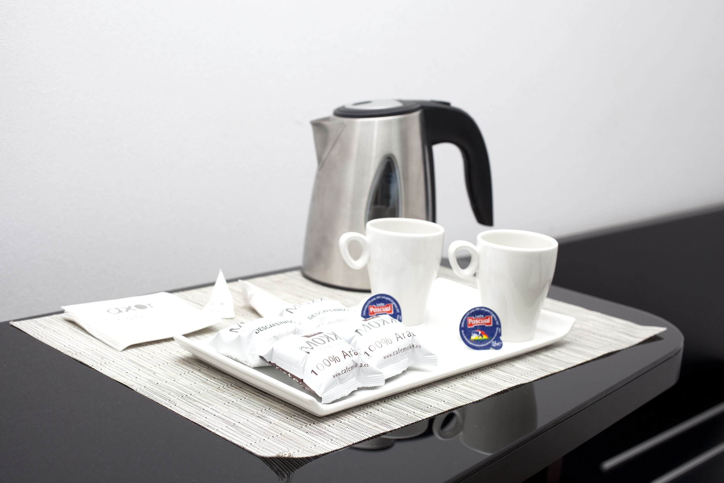 Un plateau avec deux tasses blanches, une bouilloire en argent et des sachets de café, le tout posé sur une table noire.