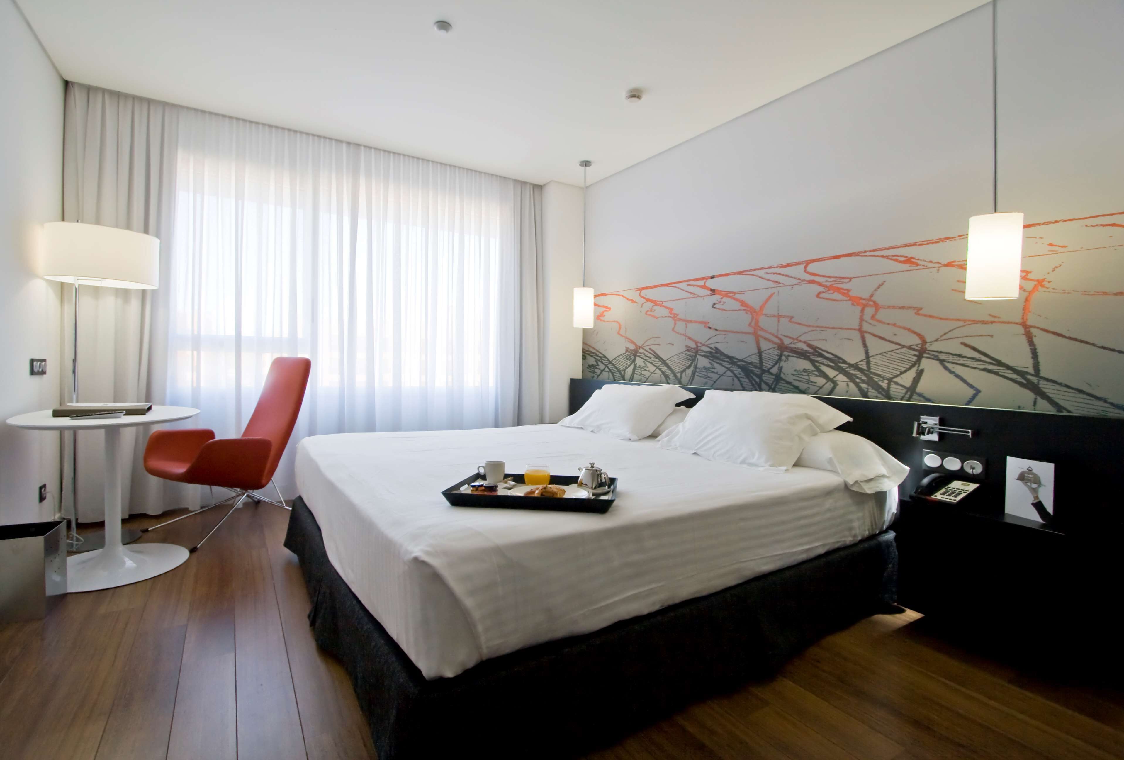 A hotel room with a bed, a desk, a chair, a tray with breakfast, and a wall with abstract art.