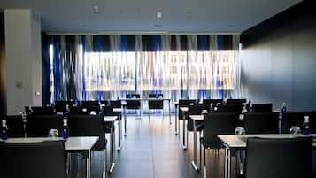 Un salón de conferencias con filas de mesas y sillas, ventanas amplias con cortinas y suelo de baldosas.