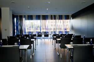 A conference room with rows of tables and chairs, large windows with curtains, and a tiled floor.