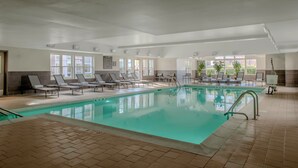 Une piscine intérieure dotée de chaises longues et de transats, entourée de fenêtres et de plantes en pot.