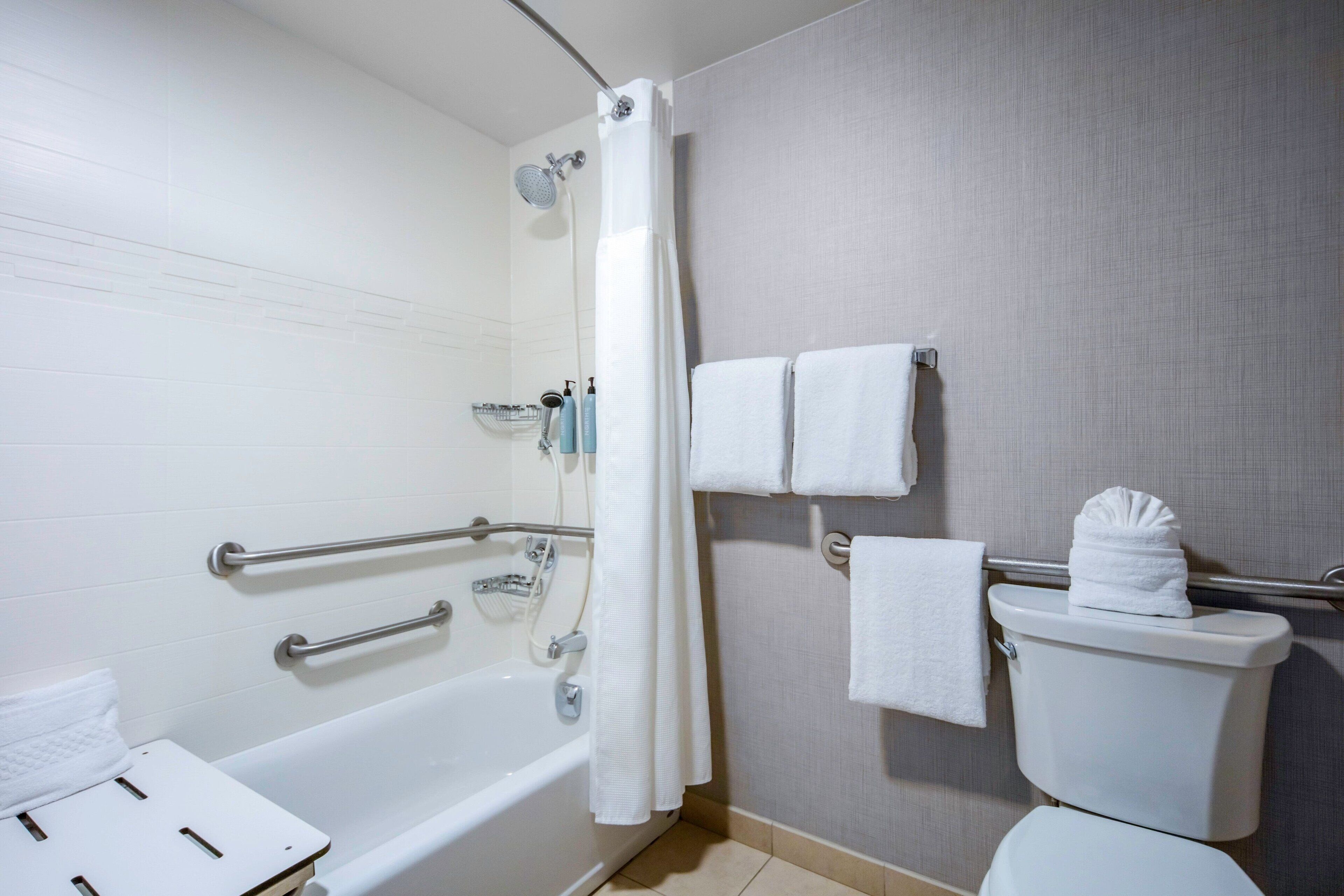 Une salle de bain équipée d’une baignoire, d’une douche et de toilettes. On y trouve des barres d’appui, un porte-serviettes et un rideau de douche.