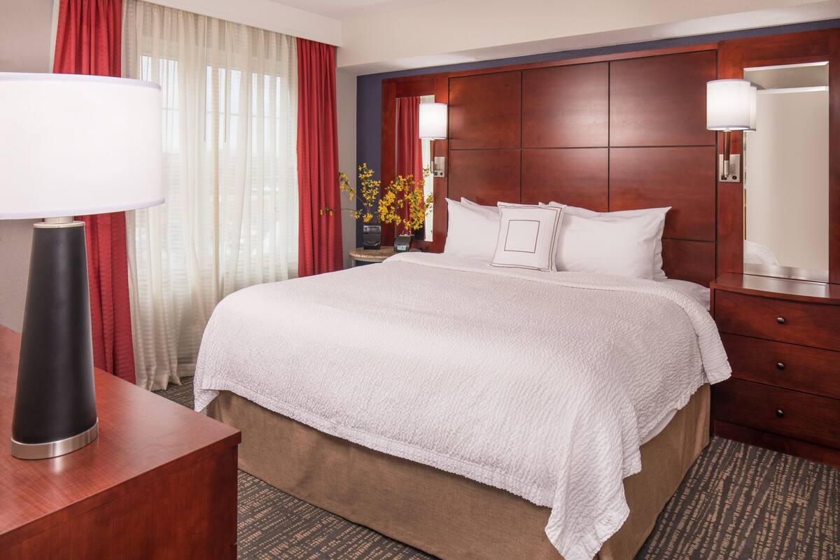 A hotel room with a large bed, a wooden headboard, a nightstand with a lamp, a dresser, and a mirror.
