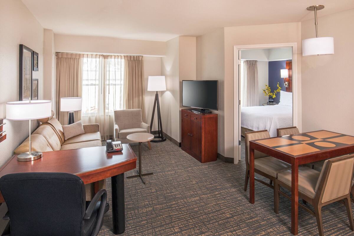 A hotel room with a bed, a desk with a chair, a TV, a lamp, a small table, and a carpeted floor.