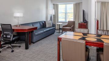 Habitación de hotel con sofá, una mesa de comedor con sillas, una mesita redonda con lámpara, televisor en un mueble bajo y una ventana con cortinas.