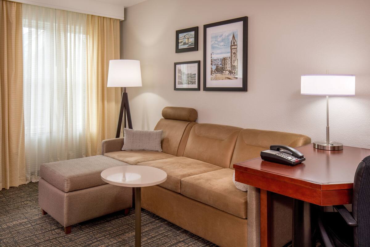 A hotel room with a sofa, a small table, a desk with a lamp, and framed pictures on the wall.