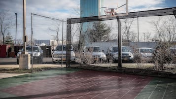 An outdoor basketball court with a net, surrounded by a fence, and parked cars in the background.