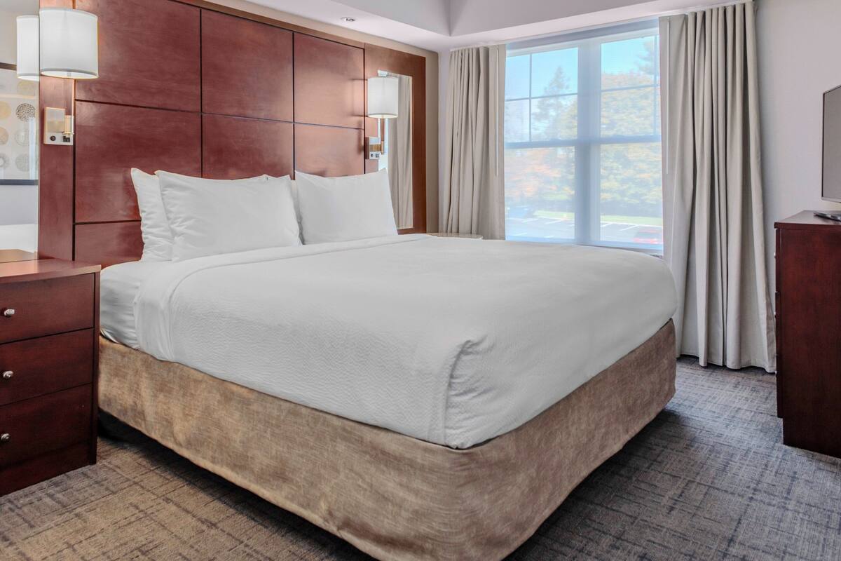 A hotel room with a large bed, wooden headboard, bedside lamps, a wooden dresser, and a large window with curtains.