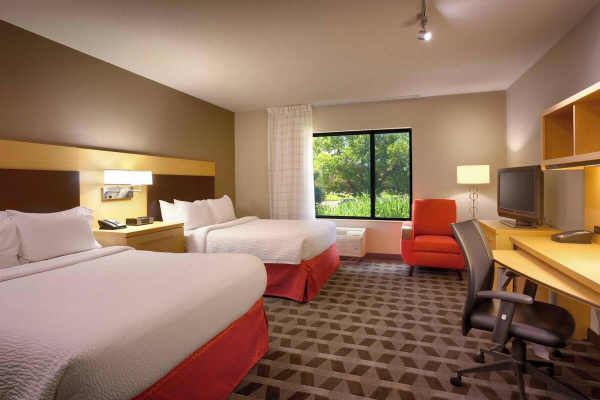 A hotel room with two beds, a desk with a computer, a chair, a TV, and a window with a view of greenery.