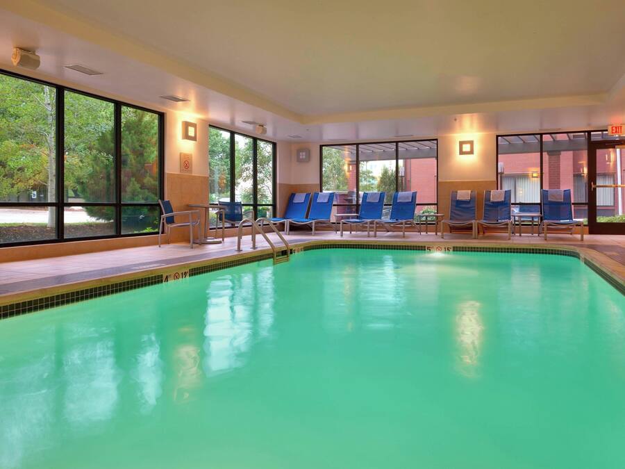 Una piscina con agua azul cristalina, rodeada de sillas de descanso y grandes ventanales que ofrecen vistas a árboles y un edificio.