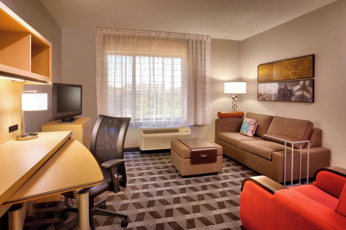 A hotel room with a desk, chair, sofa, and a small table. There is a window with curtains, a flat-screen TV, and a lamp.