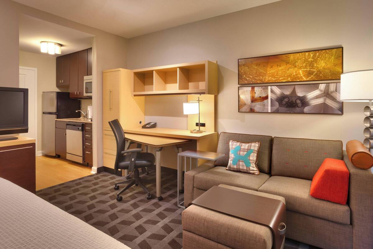A hotel room with a bed, a sofa, a desk, a chair, a television, a microwave, a refrigerator, and a kitchen area.