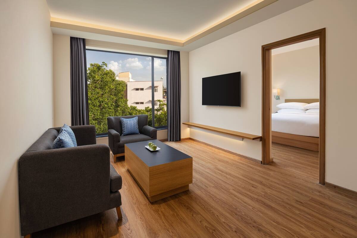 A modern hotel room with a sofa, a coffee table, a TV, and a large window offering a city view.