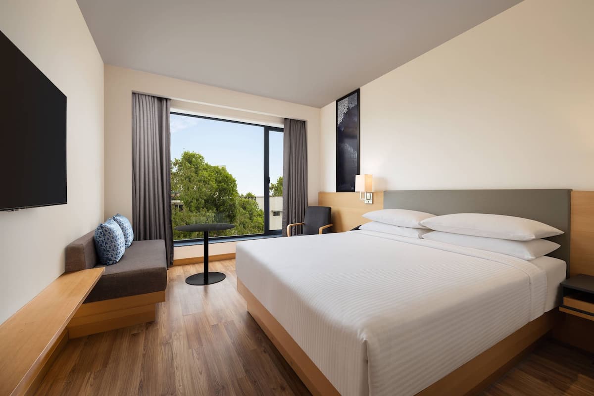 A modern hotel room with a large bed, a seating area with a view, and a flat-screen TV mounted on the wall.