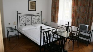 A bedroom with a metal bed frame, a bedside table with a lamp, a small table with chairs, and a television on a stand. - Hotel Terminus Vienna (Vienna)
