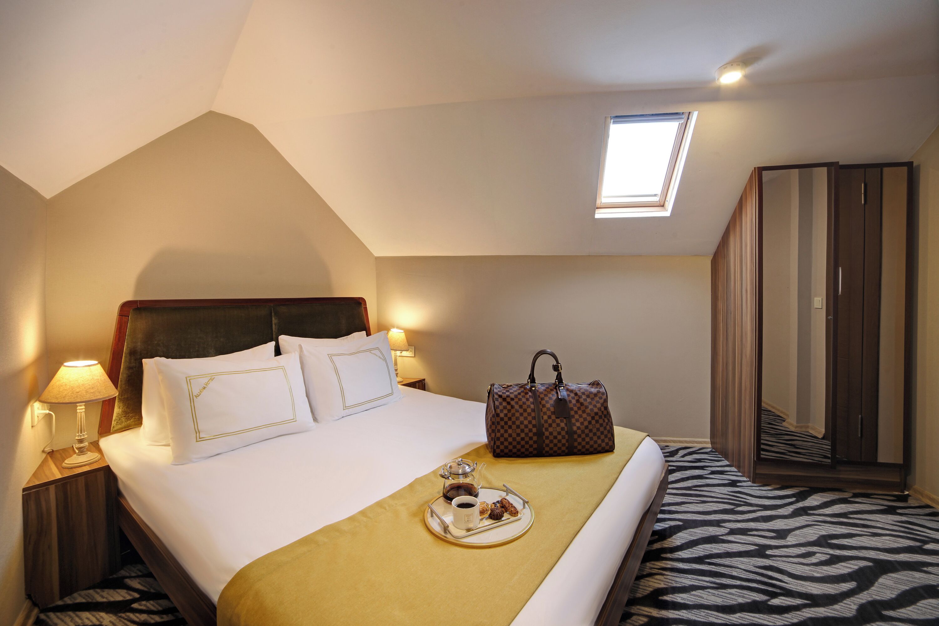 A hotel room with a bed, a bedside table with a lamp, a tray with food and a cup, a suitcase, and a wardrobe.