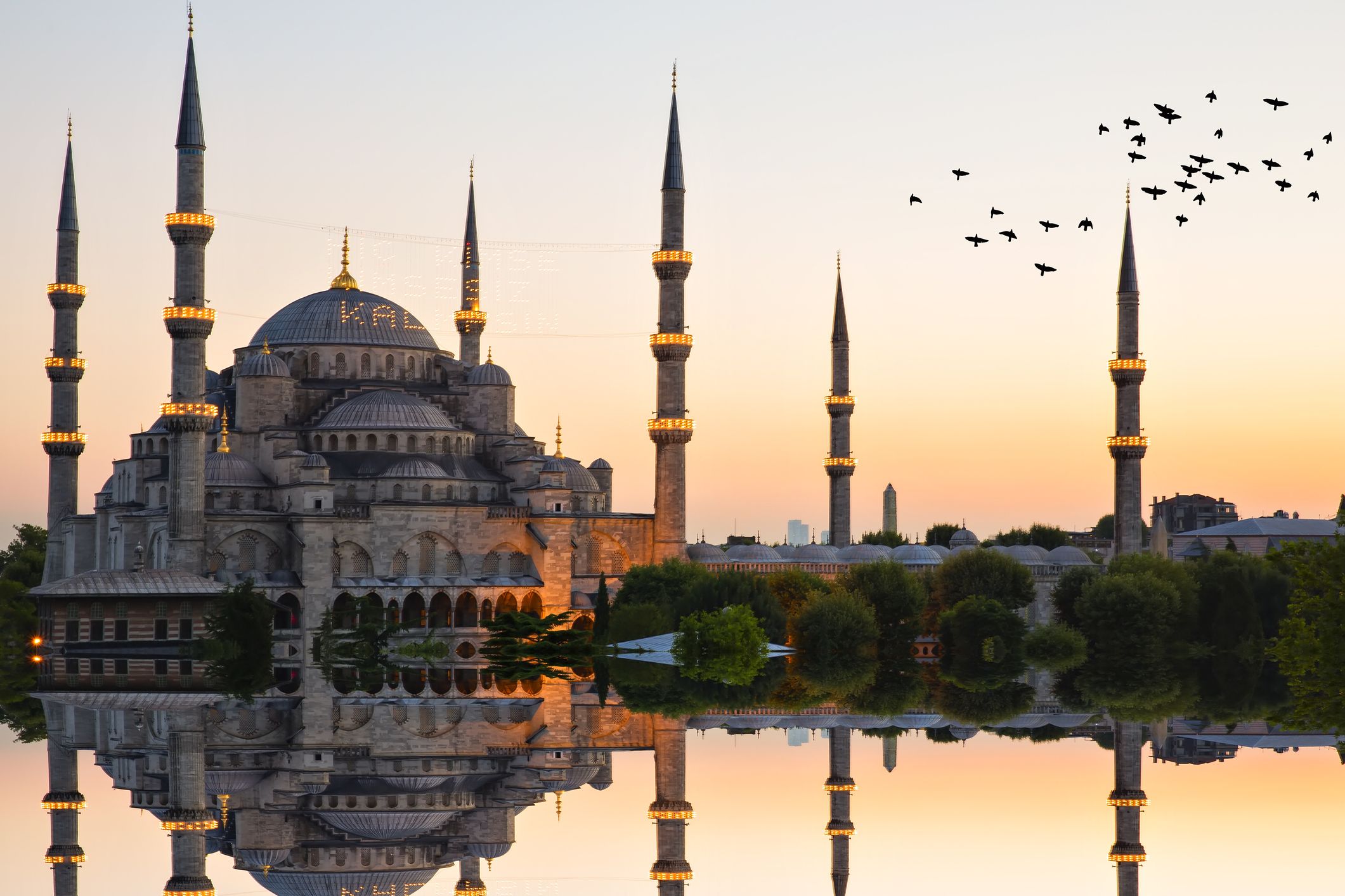 Useita minareetteja ja kupolia omaava moskeija heijastuu vesistöstä, ja taivaalla lentää lintuja.