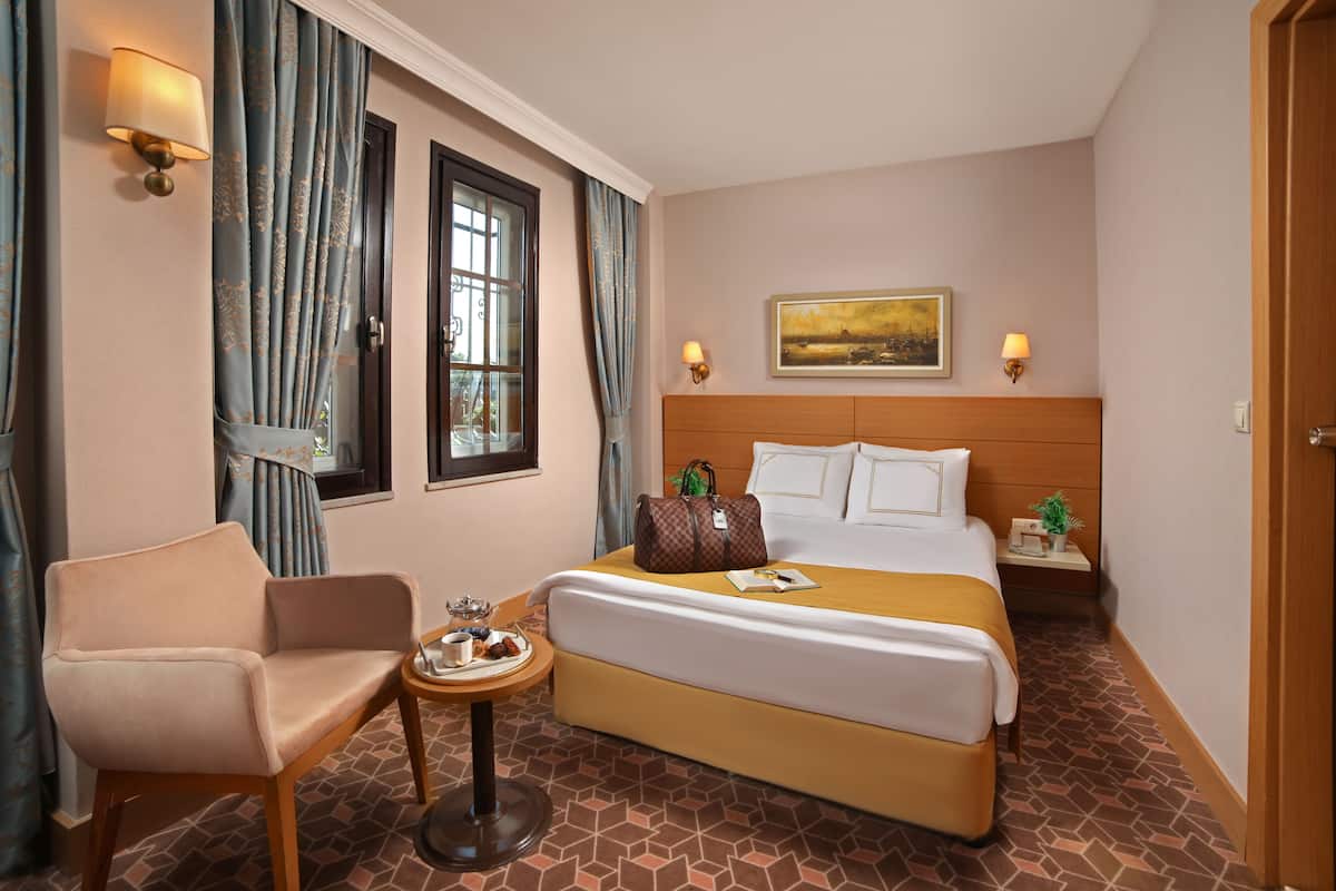 A hotel room with a bed, a chair, a small table with a tray of food, and a painting on the wall.