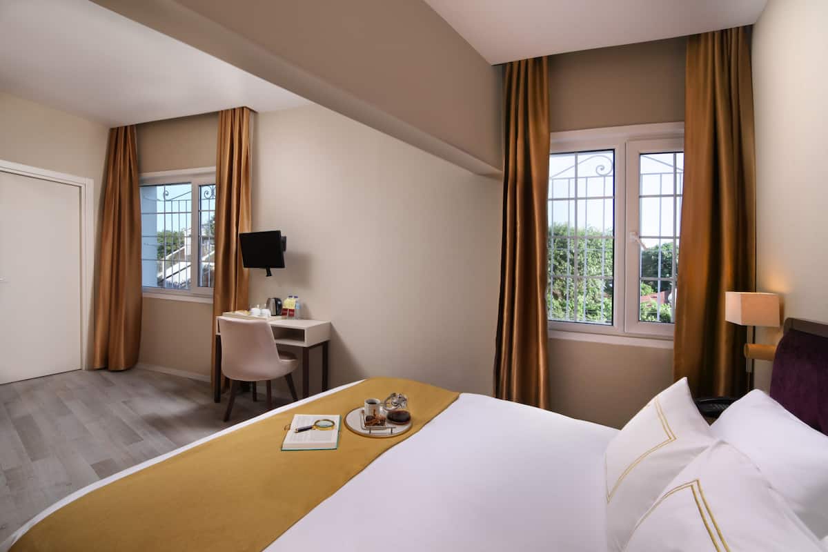A hotel room with a bed, a desk, and a chair. There is a window with curtains, a television on the wall, and a lamp on the side table.