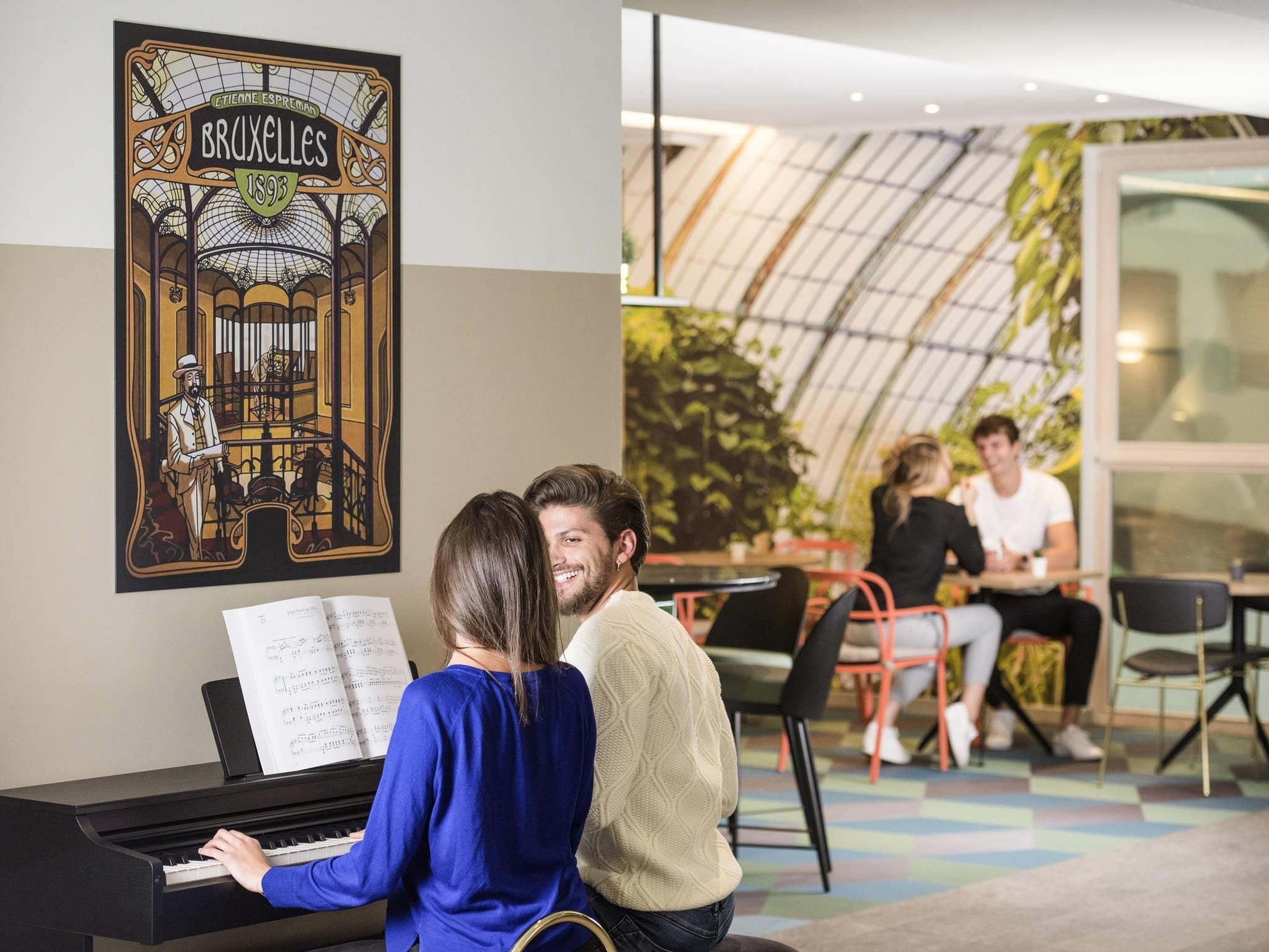 Un piano con partituras, un cartel de estación de tren y un grupo de personas en un interior moderno con techo de cristal.
