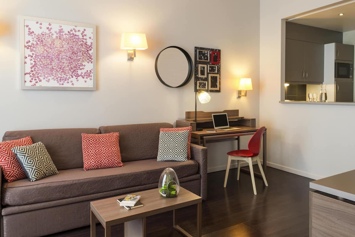 A modern living room with a sofa, a desk with a laptop, a coffee table with a book and a glass dome, and wall-mounted lamps.
