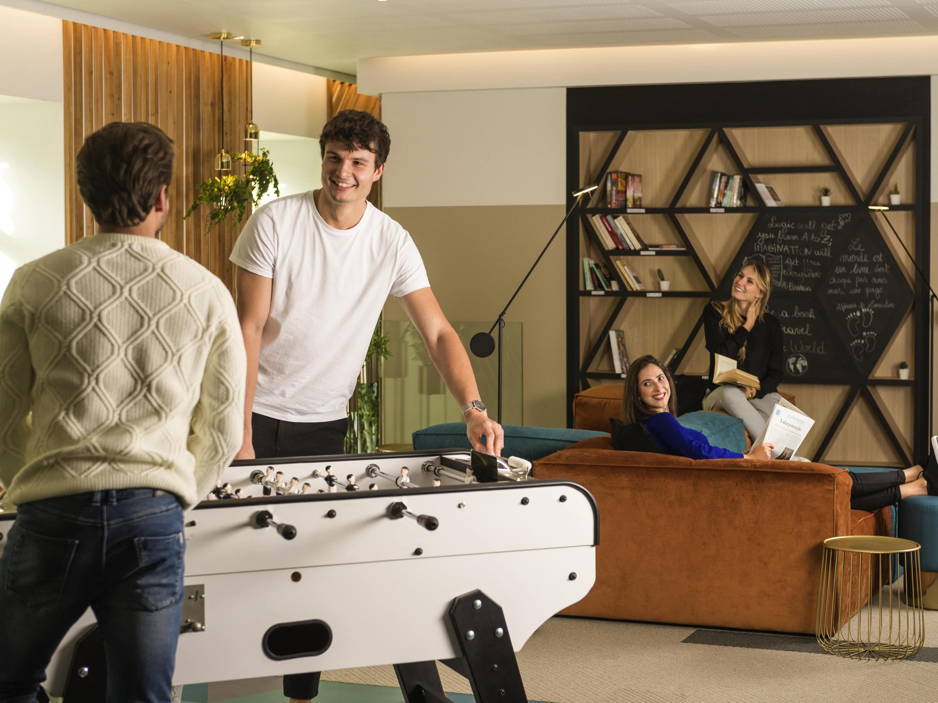 A room with a foosball table, a sofa, and a bookshelf with books and a chalkboard.