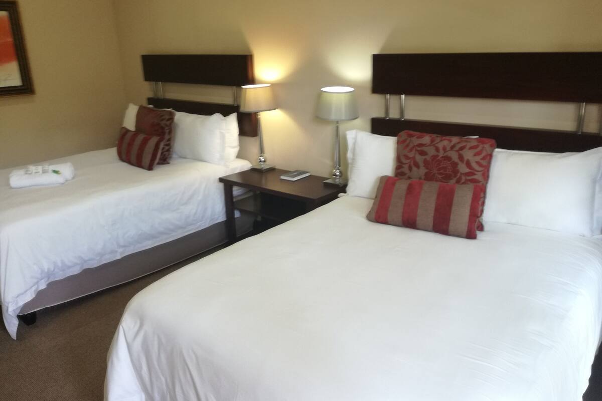 Ein Hotelzimmer mit zwei Einzelbetten, jeweils mit weißer Bettwäsche und roten und beigen Kissen. Zwei Nachttische sind vorhanden, einer mit einer Lampe und der andere mit einem Telefon.