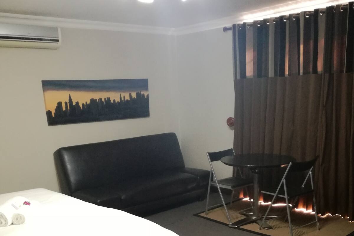 Ein Hotelzimmer mit Bett, einer schwarzen Ledersofa, einem kleinen Tisch mit zwei Stühlen, einem Bild an der Wand und einem Fenster mit Vorhängen.