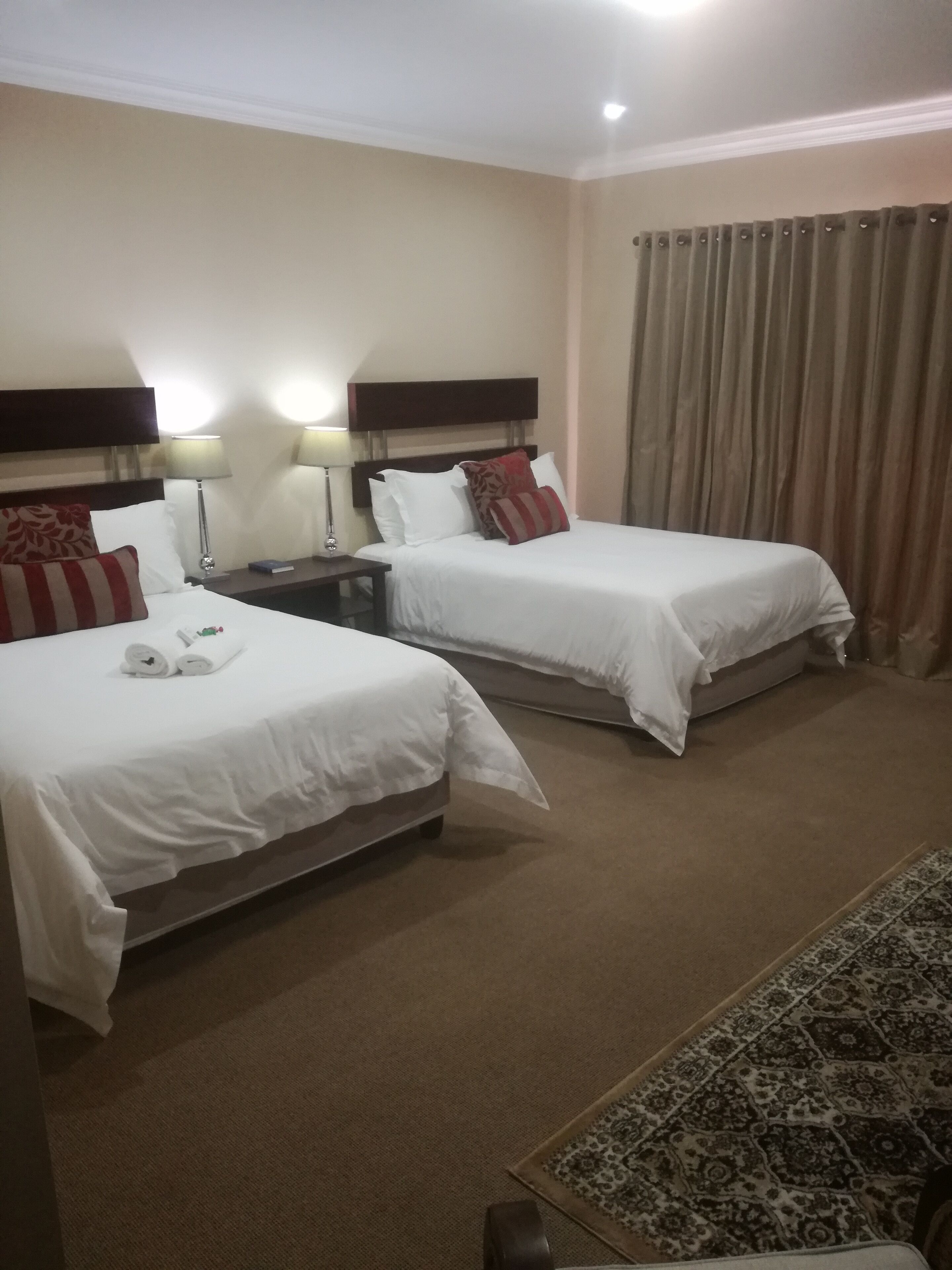 Habitación de hotel con dos camas, cada una con ropa blanca y almohadas rojas, una mesita de noche con una lámpara y una alfombra estampada en el suelo.