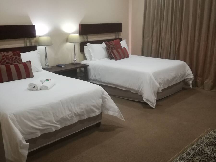 Habitación de hotel con dos camas, cada una con ropa blanca y almohadas rojas, una mesita de noche con una lámpara y una alfombra estampada en el suelo.