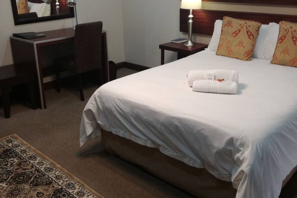 Ein Hotelzimmer mit einem ordentlich bezogenen Bett, einem Schreibtisch mit Stuhl, einer Lampe und einem gemusterten Teppich.