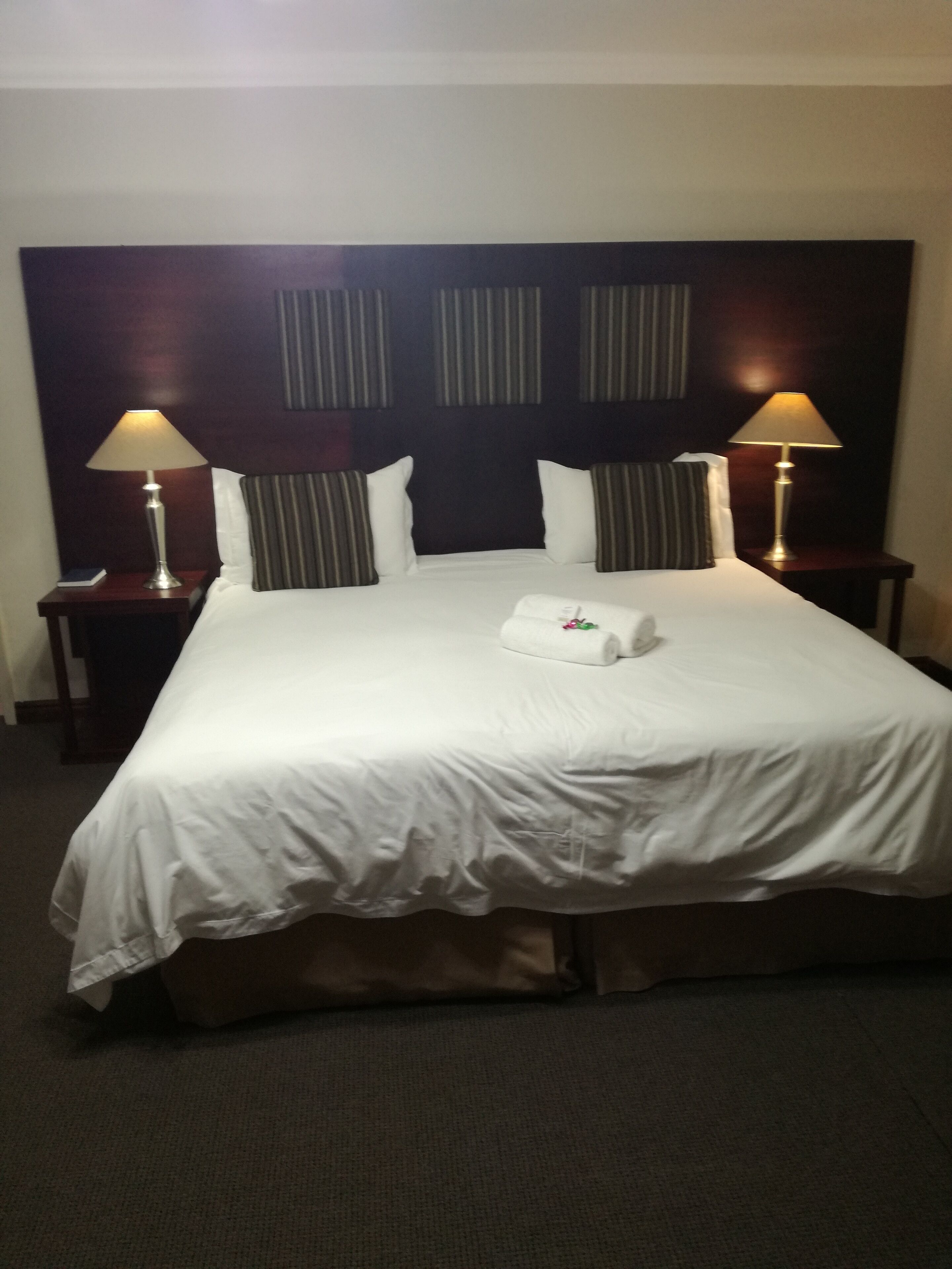 Habitación de hotel con una cama grande, dos lámparas de noche, un cabecero con rayas verticales y una mesita con un libro.