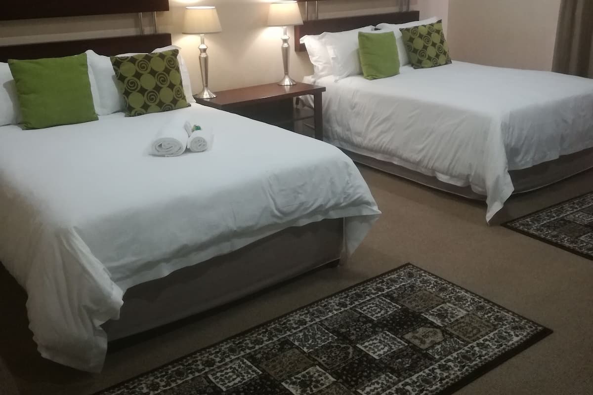 Ein Hotelzimmer mit zwei Betten, jeweils mit weißen Laken und grünen Kissen, einem Kopfteil, Nachttischlampen und einem gemusterten Teppich.