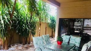 Un patio al aire libre con una mesa y sillas de vidrio, rodeado de abundante follaje y un piso de baldosas.