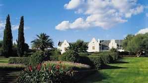 Un edificio blanco grande con múltiples chimeneas, rodeado de jardines cuidados y árboles altos.