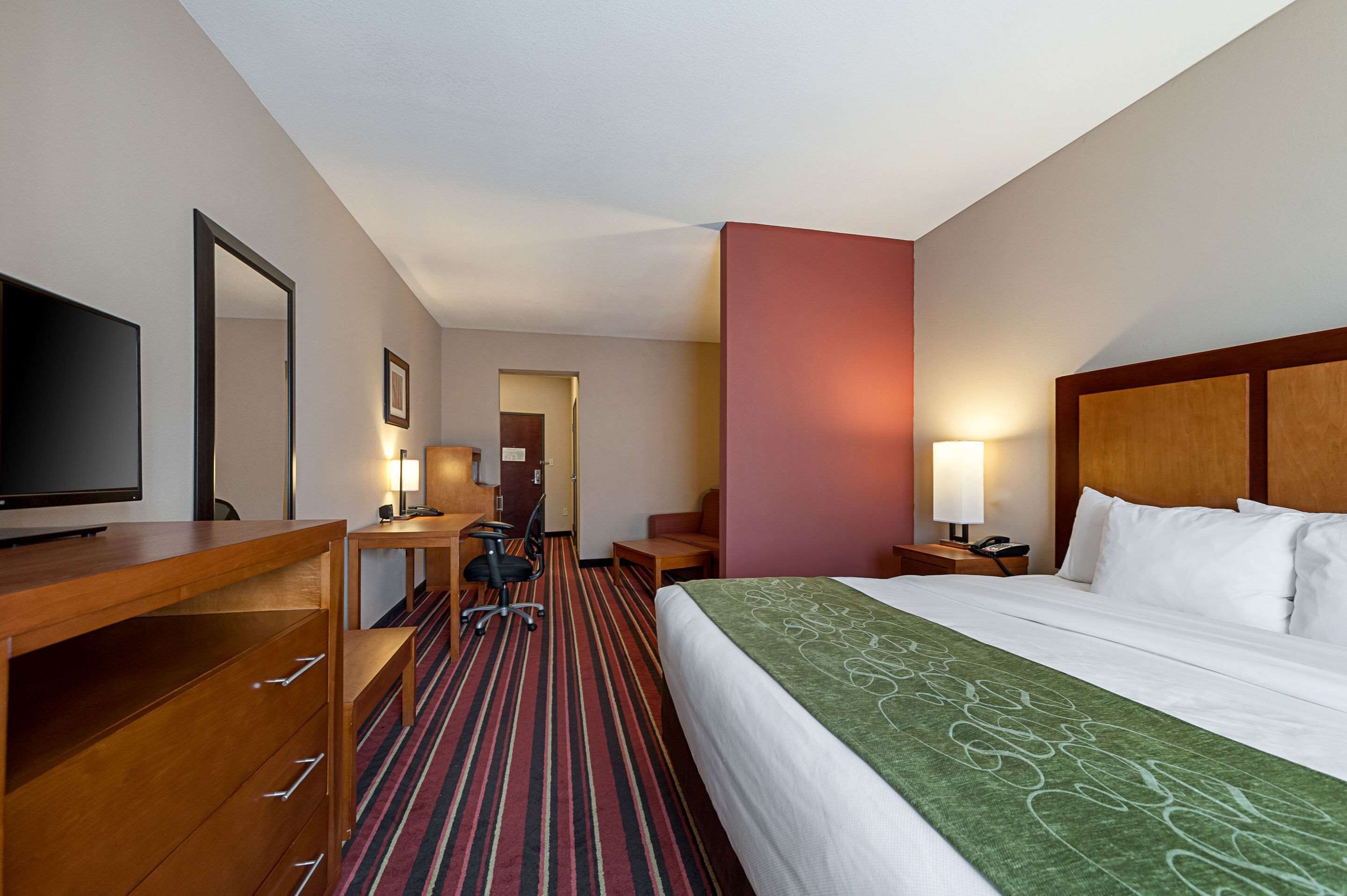 Una habitación de hotel con cama, escritorio con silla, televisión, lámpara de escritorio y mesita de noche con lámpara.